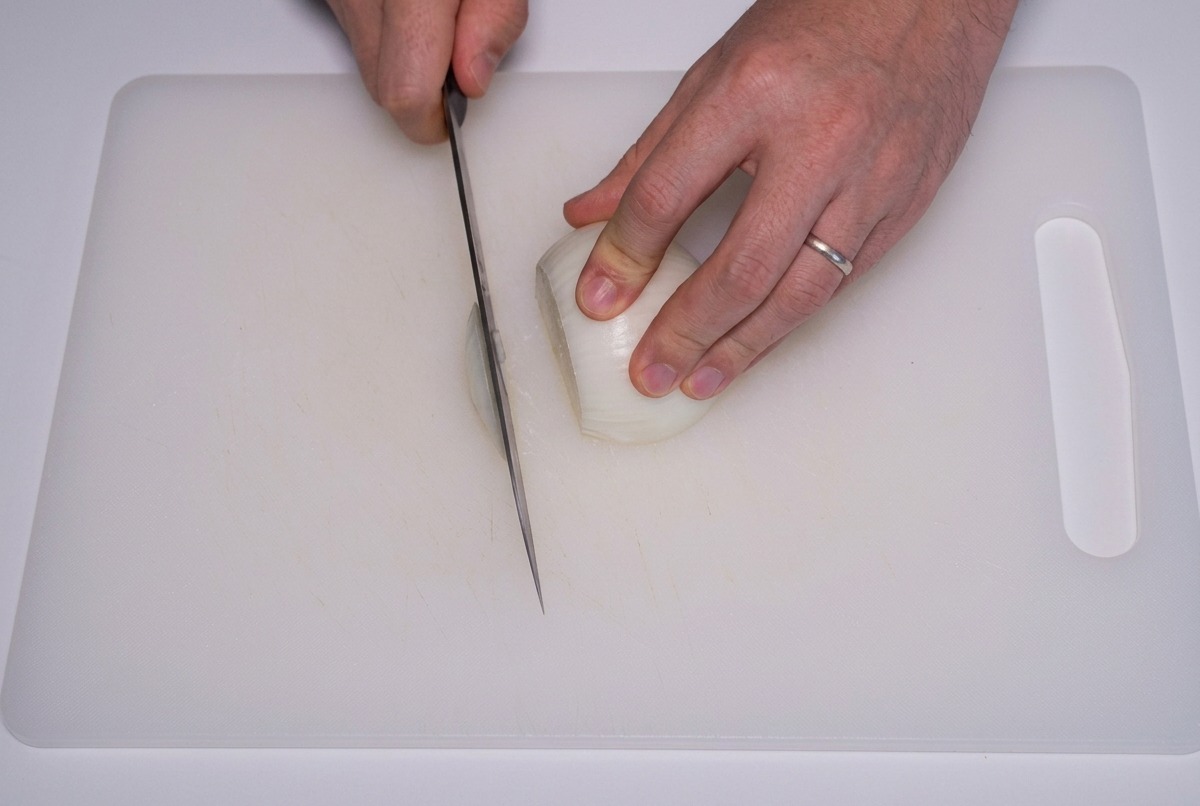 Une personne coupe un oignon blanc en fines lamelles verticales sur une planche à découper blanche.