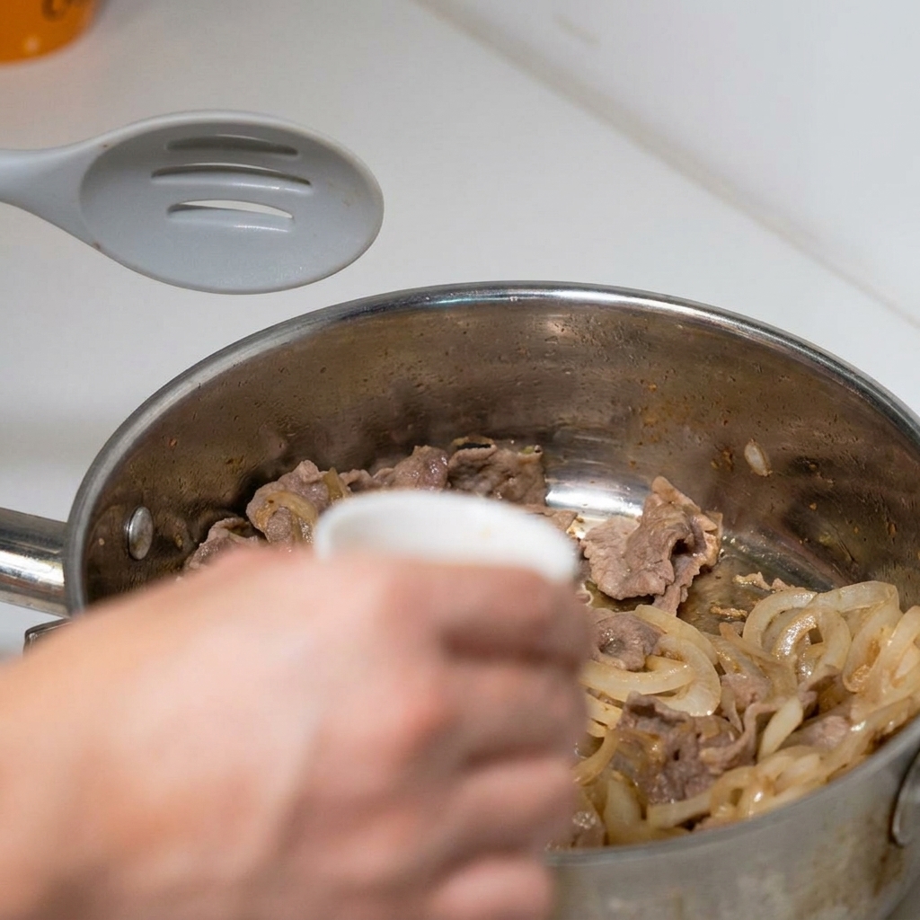 Une main verse un petit bol de sauce japonaise sur le mélange de bœuf et doignons en cours de cuisson.