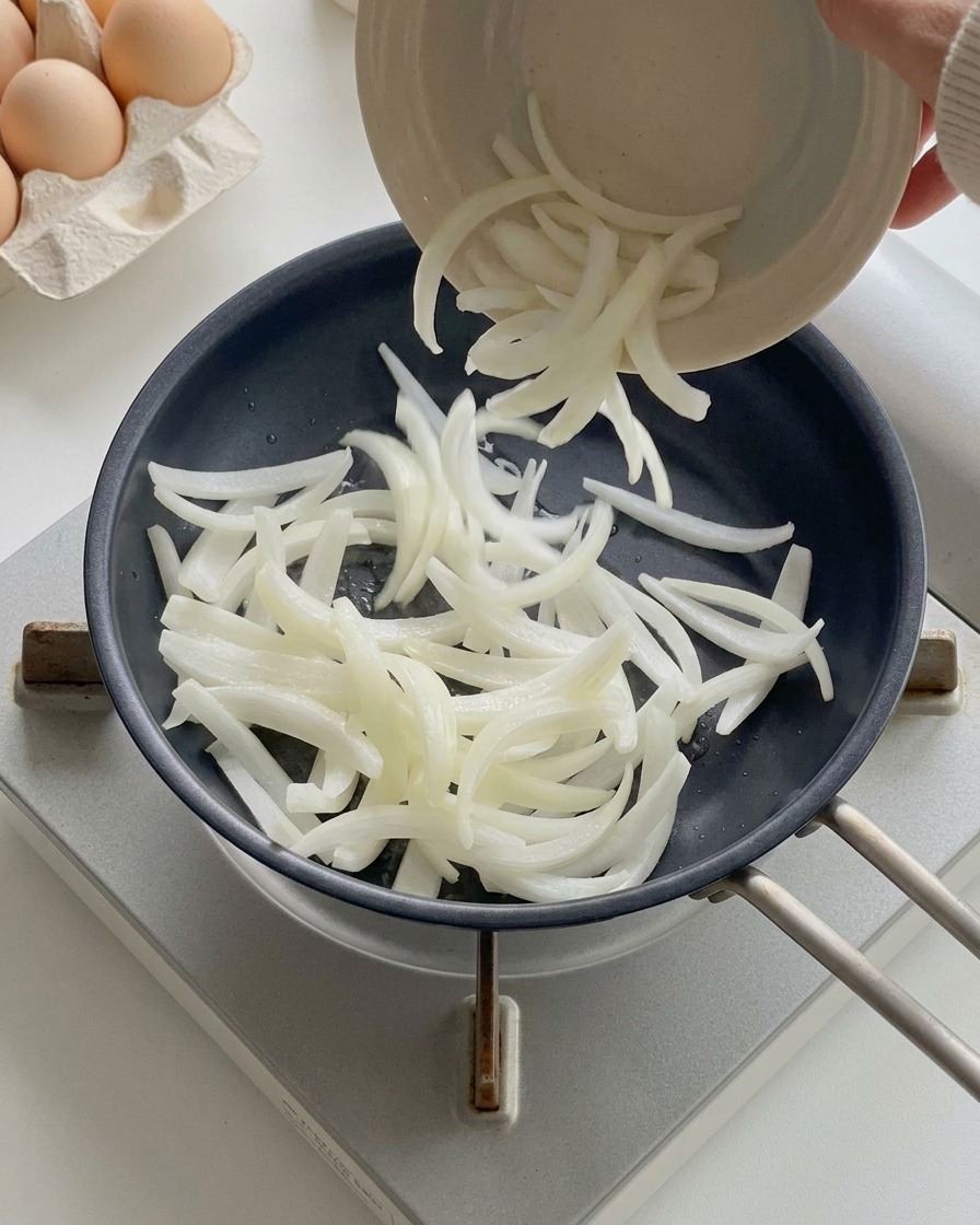 Oignons blancs émincés versés depuis un petit bol dans une poêle noire chaude.