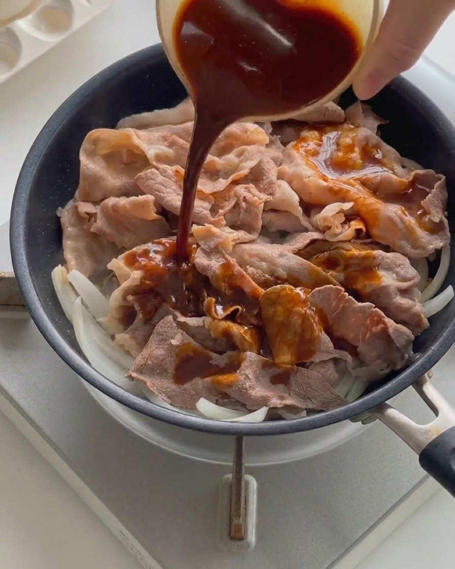Sauce liquide foncée versée depuis un petit bol sur les tranches de bœuf et les oignons dans une poêle chaude.