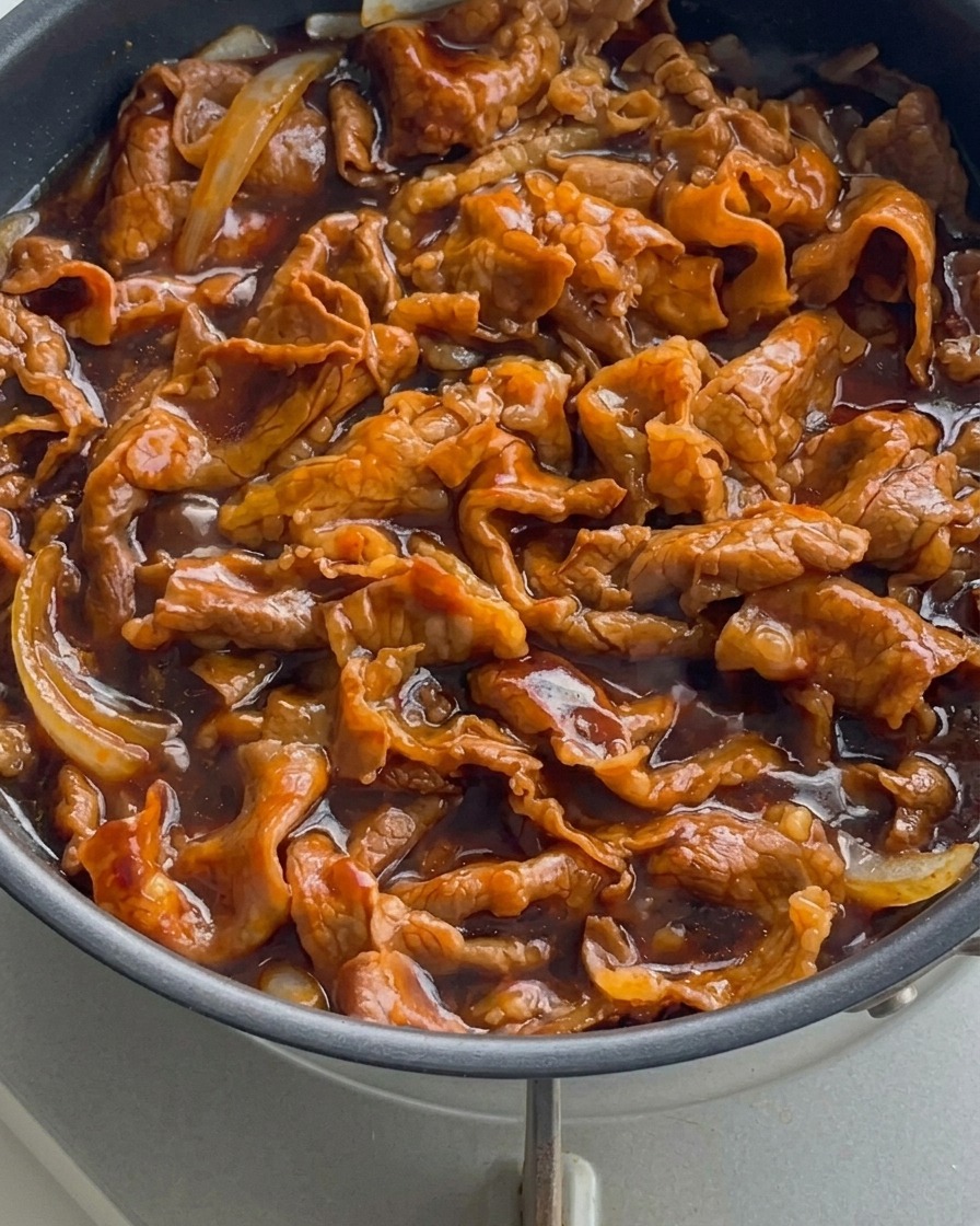 Gros plan de fines tranches de bœuf et doignons bouillonnant dans une sauce épaisse, sombre et brillante.