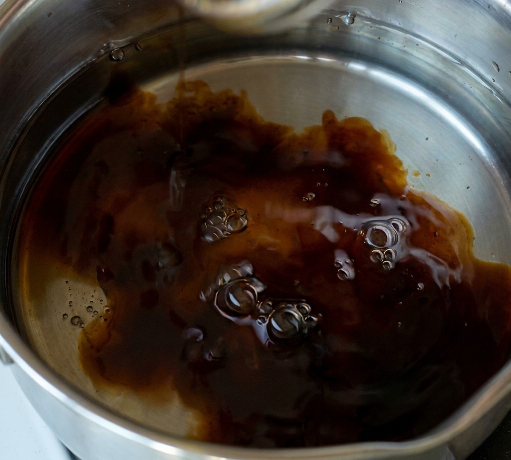 Une main ajoutant une cuillerée de sucre blanc en poudre dans une casserole en acier inoxydable contenant une sauce sombre frémissante à base de soja.