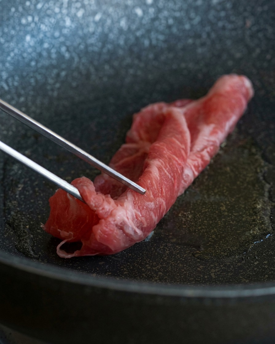 Une pince en métal déposant une fine tranche de bœuf cru persillé dans une poêle chaude contenant de lhuile scintillante.