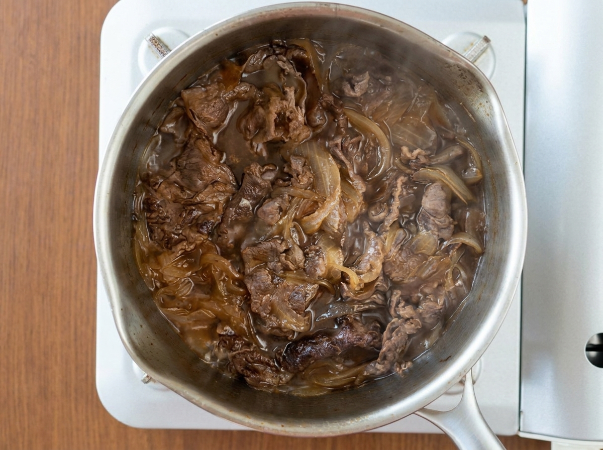 Vue de dessus de tranches de bœuf et doignons mijotant ensemble dans une sauce sombre et bouillonnante à lintérieur dune casserole en acier inoxydable.