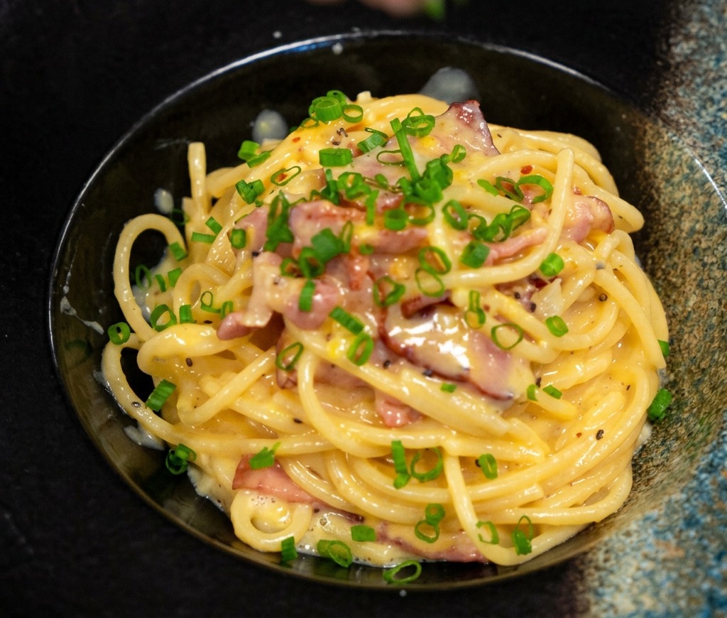Doigts saupoudrant des oignons verts hachés sur un bol de pâtes crémeuses au bacon élégamment dressé.