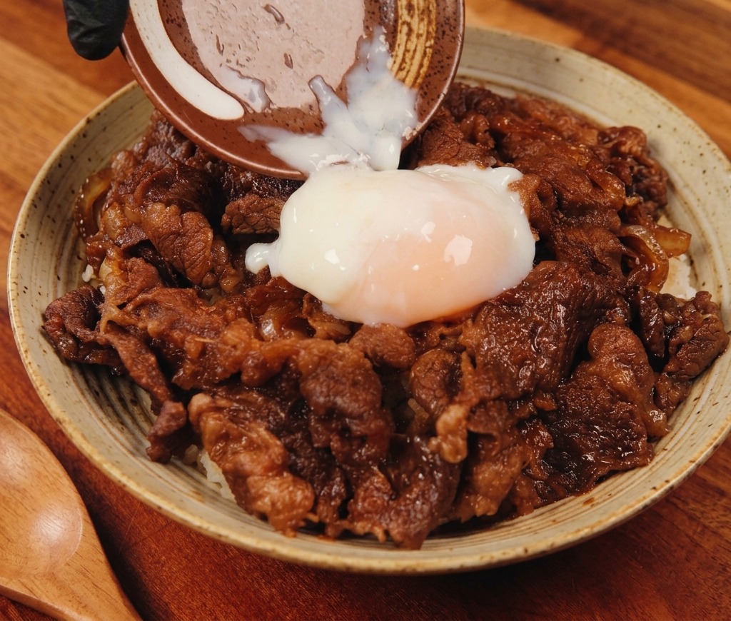 Un œuf onsen mollet est glissé depuis une petite coupelle en céramique sur un bol fumant de bœuf et de riz savoureux.