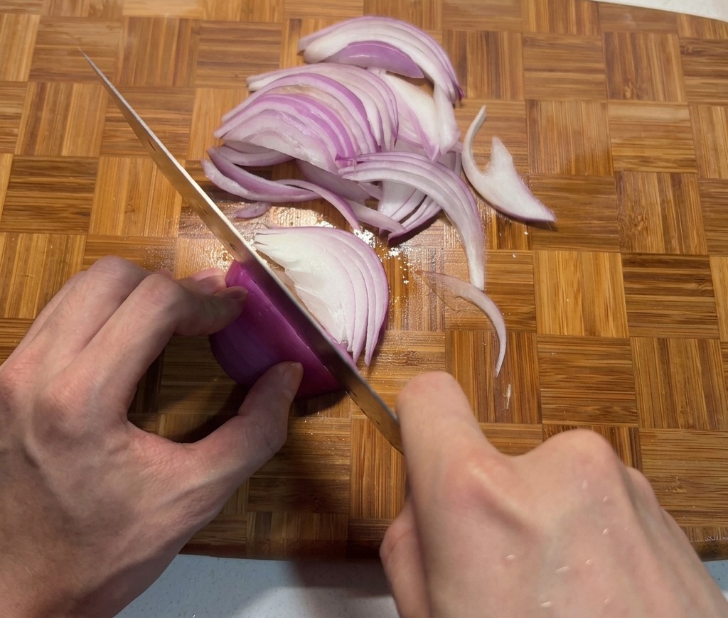 Mains éminçant un oignon rouge en fines lamelles sur une planche à découper en bambou avec un couteau de chef.
