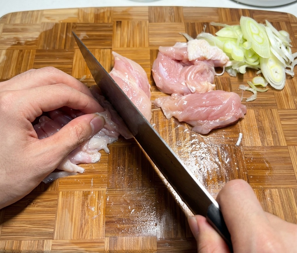 Découpe de hauts de cuisse de poulet crus en petits morceaux sur une planche en bois.