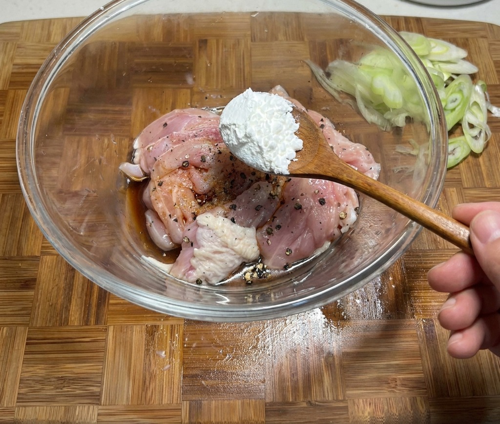 Ajout dune cuillérée de fécule de maïs dans un bol de poulet cru mariné dans de la sauce soja et des épices.