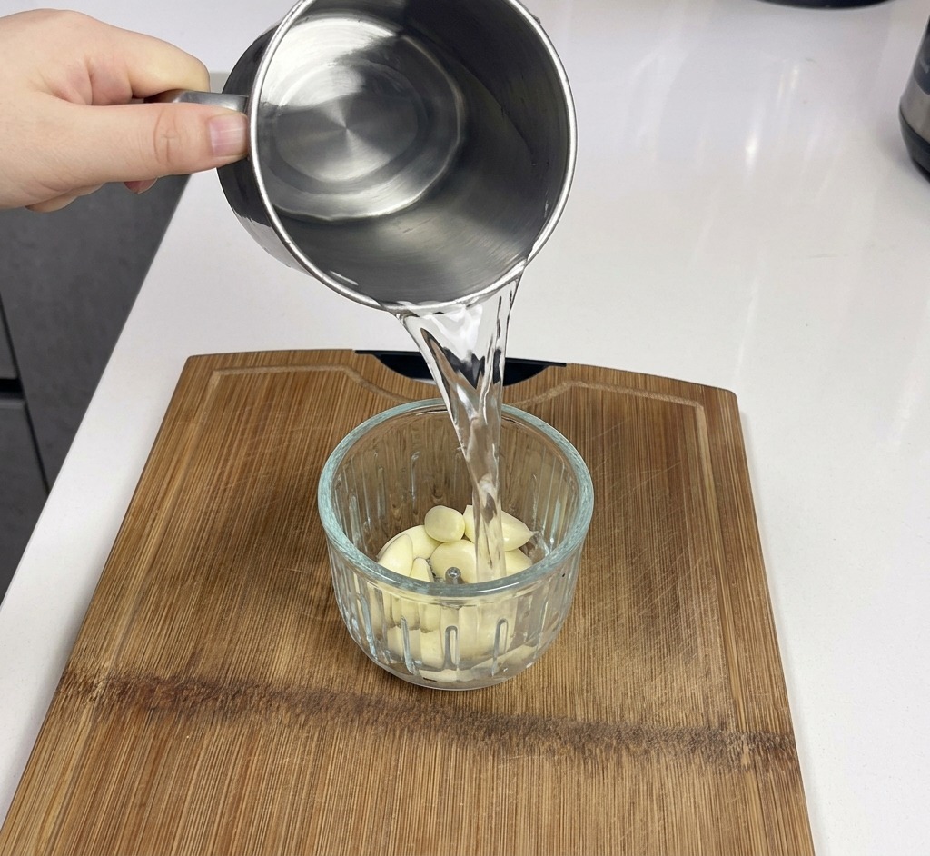 Un pichet en métal versant de leau dans un mixeur en verre contenant des gousses dail blanc frais.
