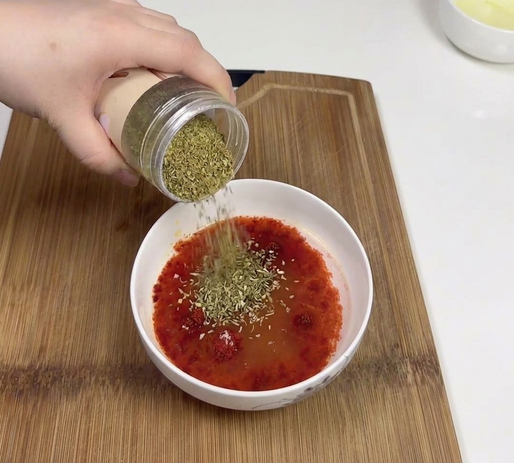 Une main versant du cumin vert en poudre depuis un bocal dans un bol contenant une base de sauce au piment rouge.
