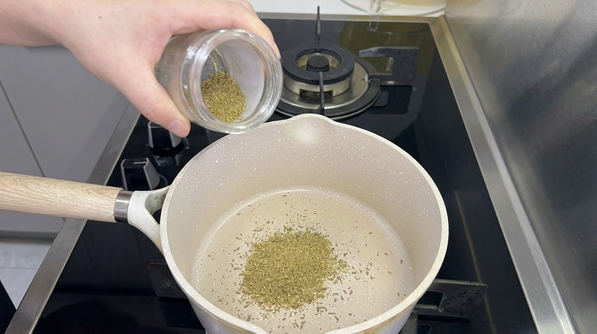 Du cumin en poudre versé depuis un bocal en verre dans une casserole blanche posée sur une cuisinière à gaz noire.