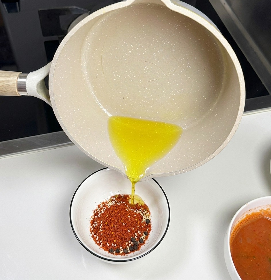Une casserole en céramique claire versant de lhuile chaude transparente dans un petit bol blanc rempli de piment rouge en poudre et de graines de sésame.