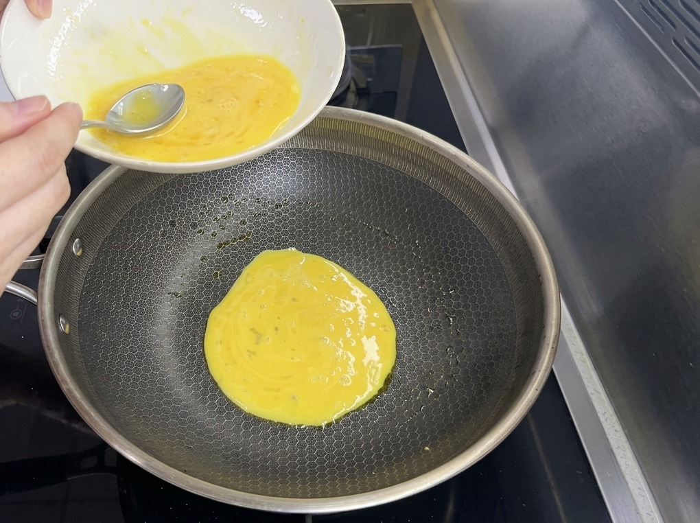 Une pâte à crêpe fine aux œufs de couleur jaune versée depuis un bol dans une grande poêle à motifs hexagonaux.