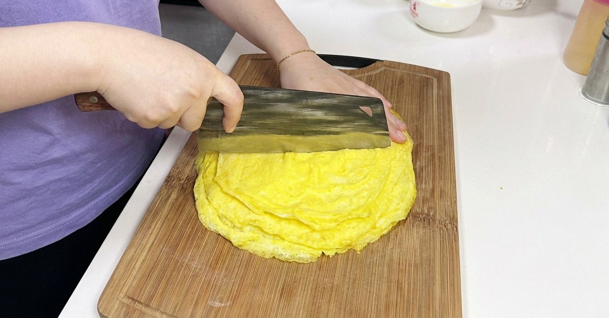 Une personne utilisant un grand couperet chinois pour couper une pile de fines crêpes aux œufs jaunes cuites sur une planche en bois.