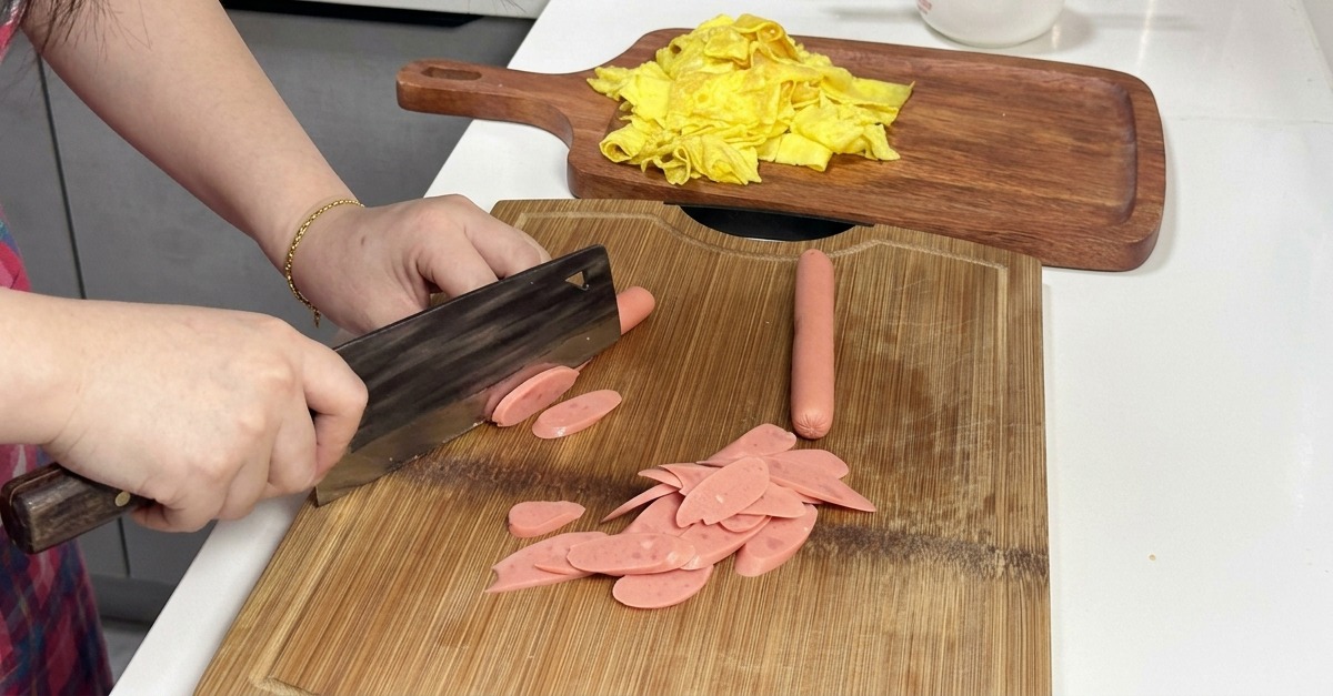 Une personne coupant de fines tranches de saucisses roses en biais sur une planche à découper en bois avec un couperet.