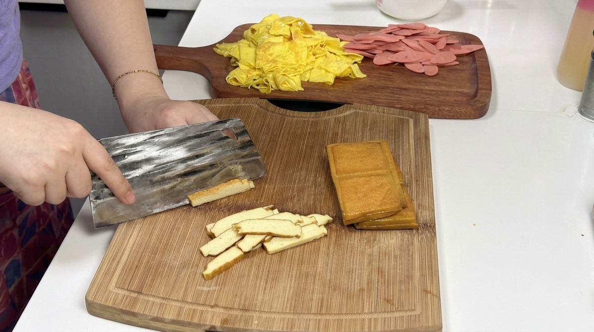 Un couperet en métal tranchant une pile de blocs de tofu fumé brun doré sur une planche à découper en bois, avec des lanières dœufs et de jambon préparées en arrière-plan.