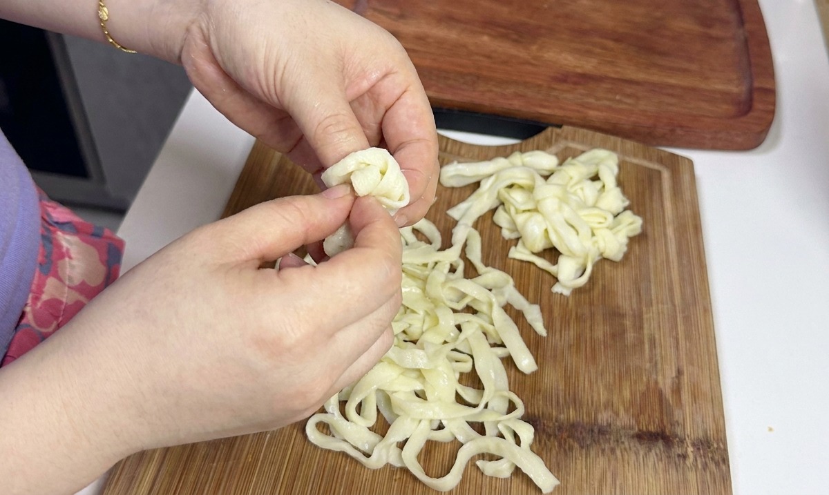 Des mains séparant et desserrant soigneusement un tas de nouilles de riz froides, larges, plates et blanches sur une planche à découper en bois.