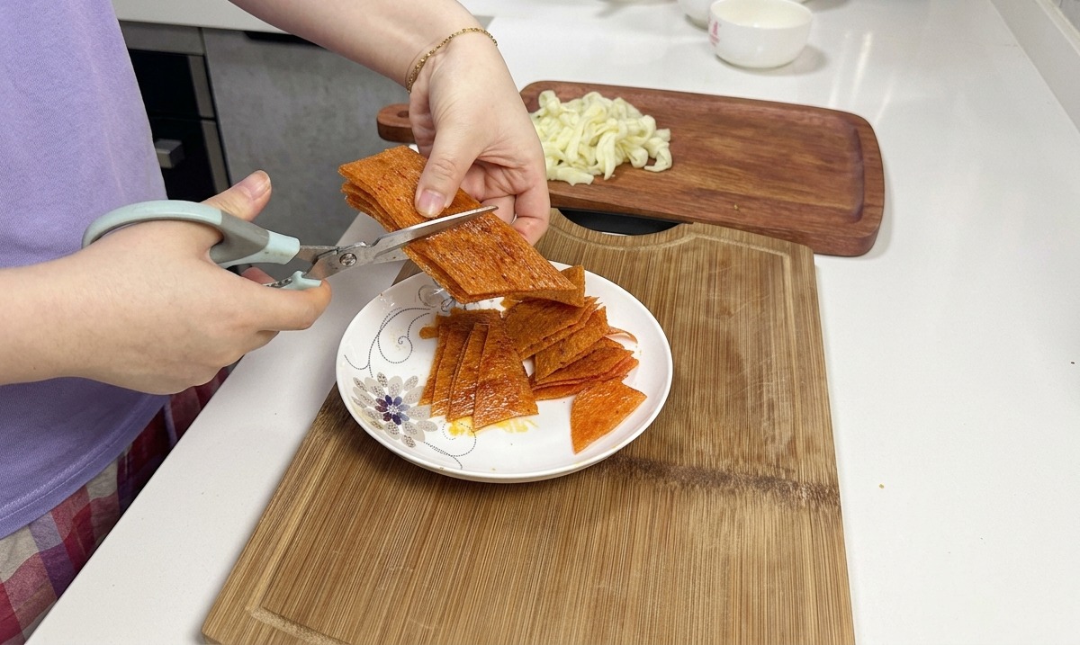 Des ciseaux de cuisine coupant une pile de lanières de latiao épicées de couleur orange au-dessus dune assiette en céramique blanche.