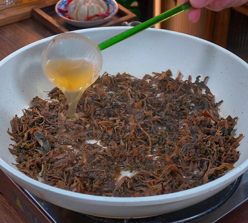 Gras liquide doré versé dune louche verte sur des feuilles de moutarde hachées et sautées dans une poêle blanche.