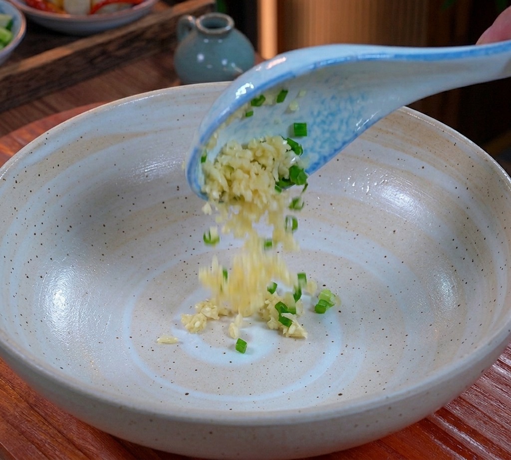 Ail émincé et oignons verts hachés tombant dune cuillère bleue et blanche au fond dun grand bol en céramique.