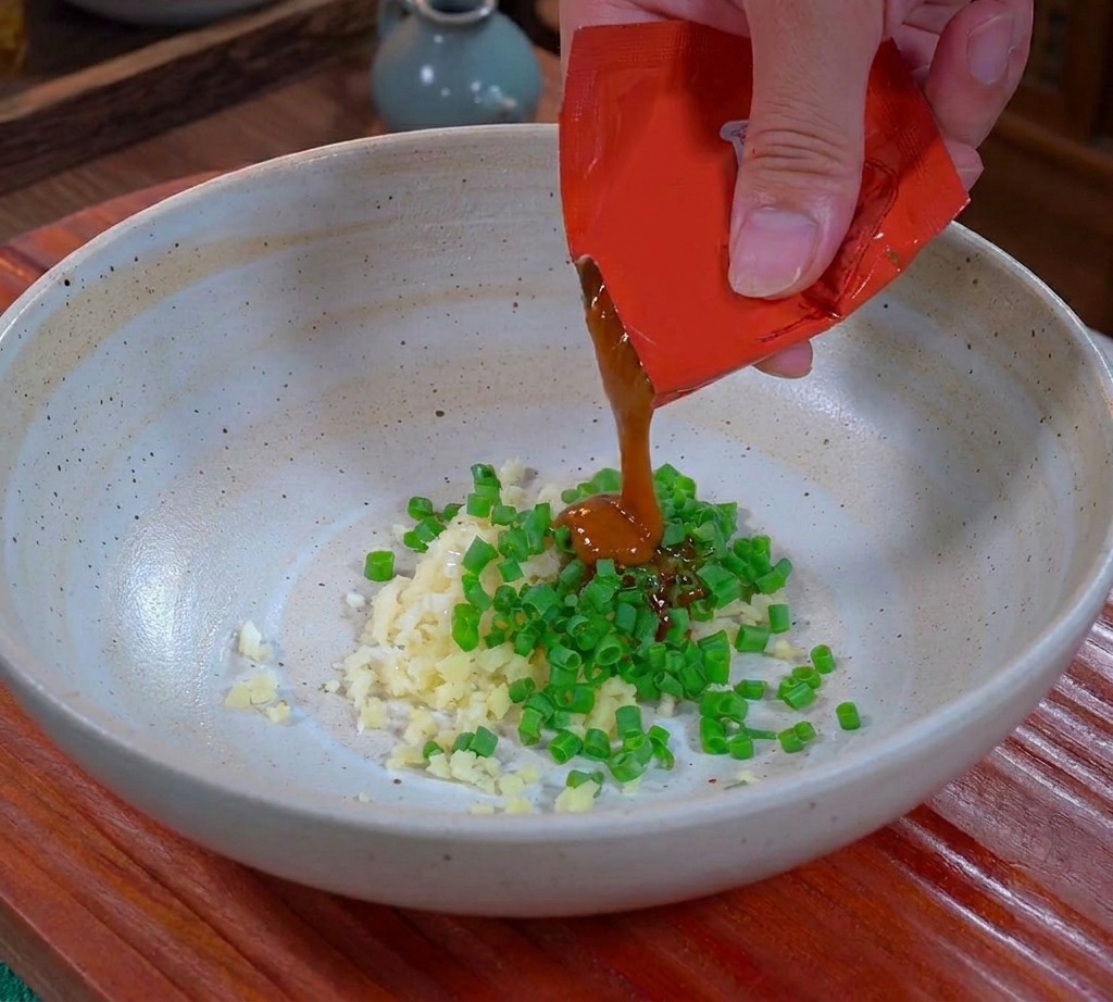 Une main pressant de la sauce Dandan marron et épaisse dun sachet rouge dans un bol en céramique sur de lail et des oignons verts hachés.