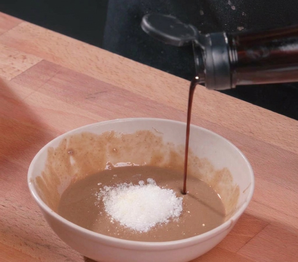 Un filet de sauce soja foncée versé dans un bol en céramique contenant de la pâte de sésame, un monticule de sucre blanc et du vinaigre.