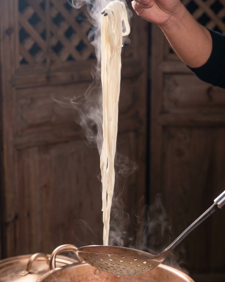 Un fagot de nouilles blanches fumantes soulevé verticalement dune marmite en cuivre à laide de baguettes et dune écumoire.