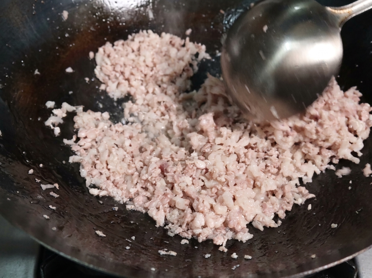 Porc haché sauté avec une louche en métal dans un wok très chaud, montrant la viande qui commence à cuire et à se détacher.