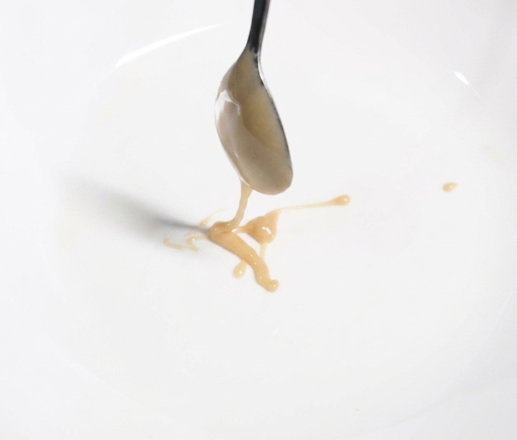 Une cuillère en métal versant une pâte de sésame épaisse et beige dans un bol blanc pour créer la sauce.