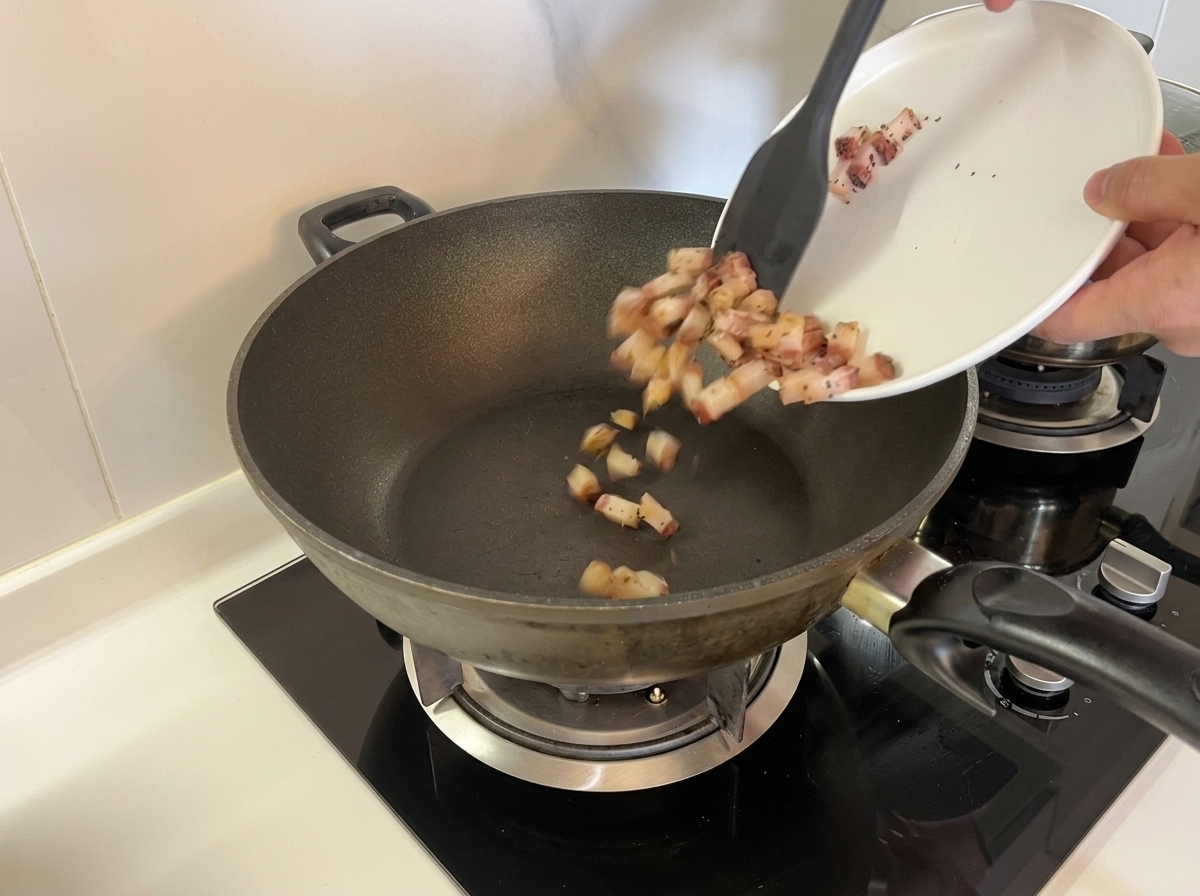 Du guanciale cru en dés versé dune assiette blanche dans un wok sec et froid à laide dune spatule.