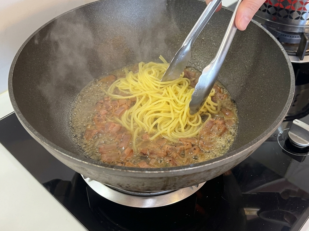 Pinces en métal transférant des spaghettis cuits dans un wok rempli de guanciale et dun peu de liquide chaud.