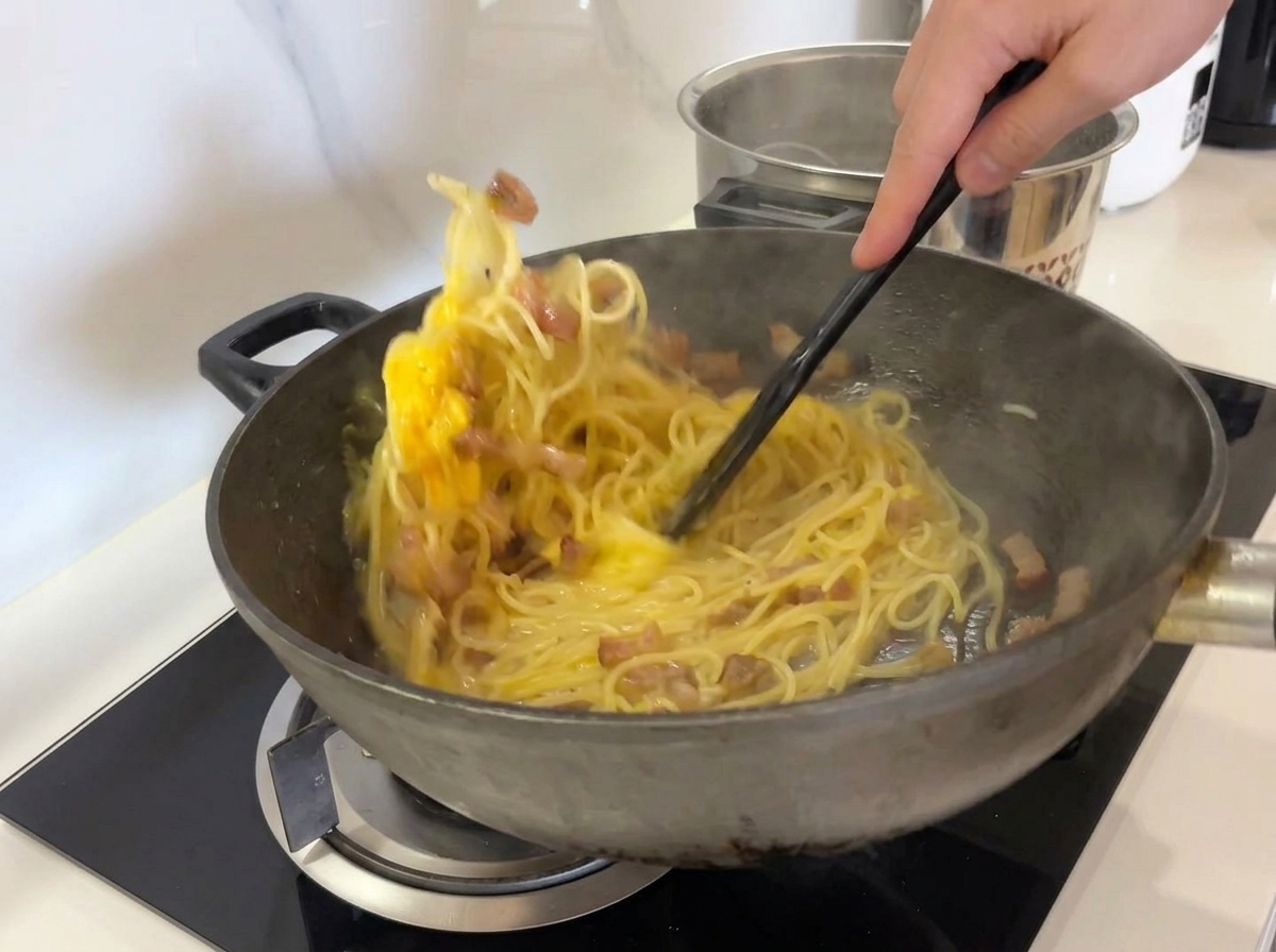 Mélanger énergiquement spaghettis, guanciale et sauce aux œufs dans un wok avec une spatule noire.