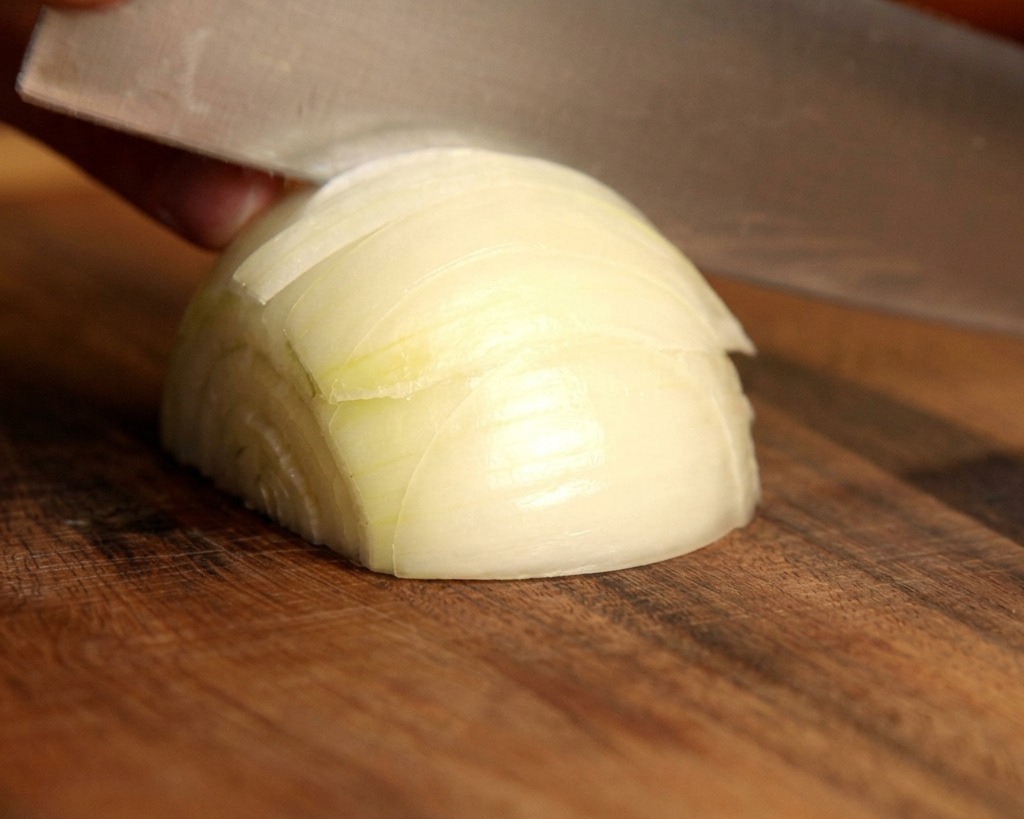 Un couteau de chef tranchant coupant un oignon blanc frais en fins quartiers sur une planche en bois.