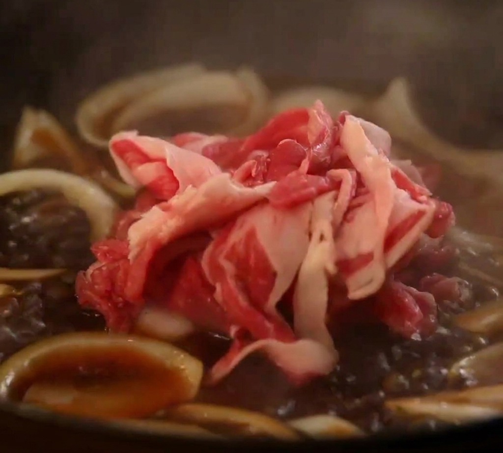 De fines tranches de bœuf cru déposées dans une poêle où mijotent des oignons dans un bouillon japonais.