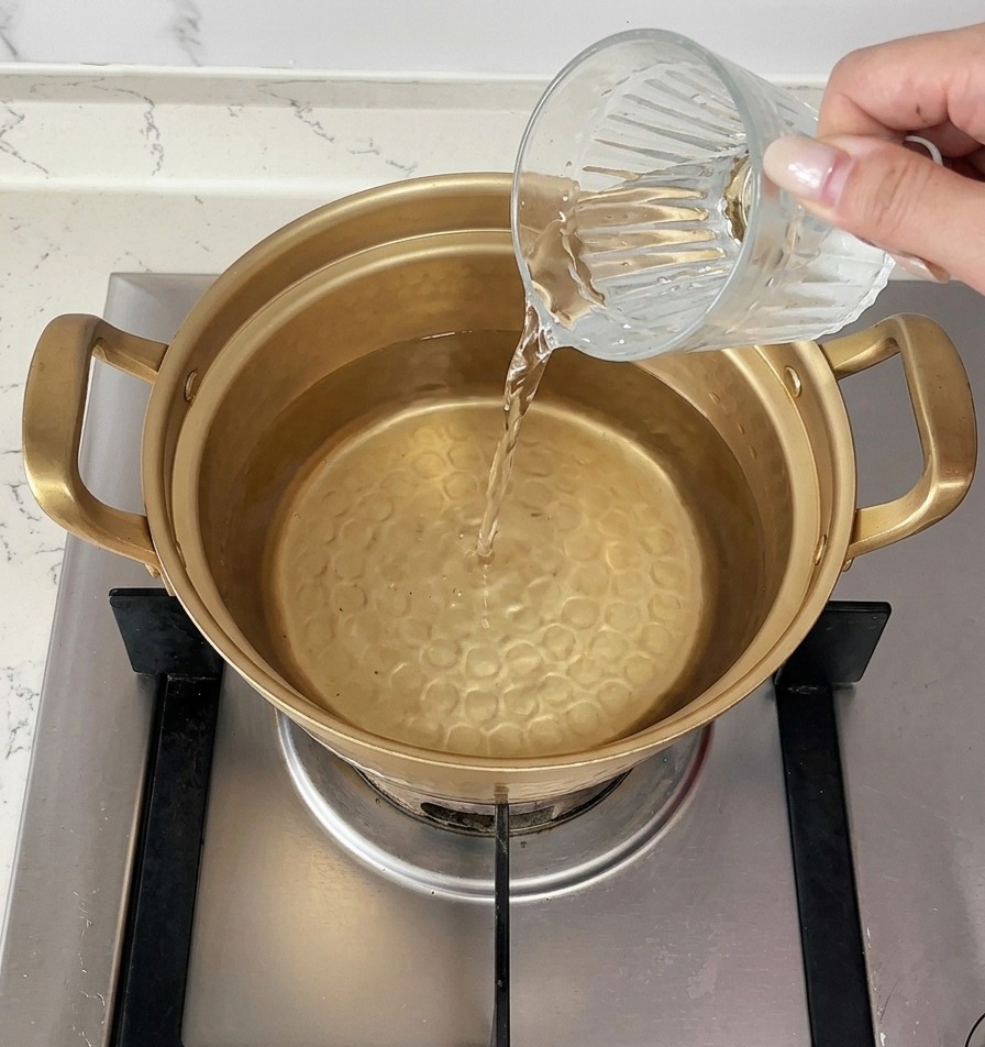 Une main versant une tasse deau froide dans une casserole dorée déjà remplie deau chaude sur la cuisinière.