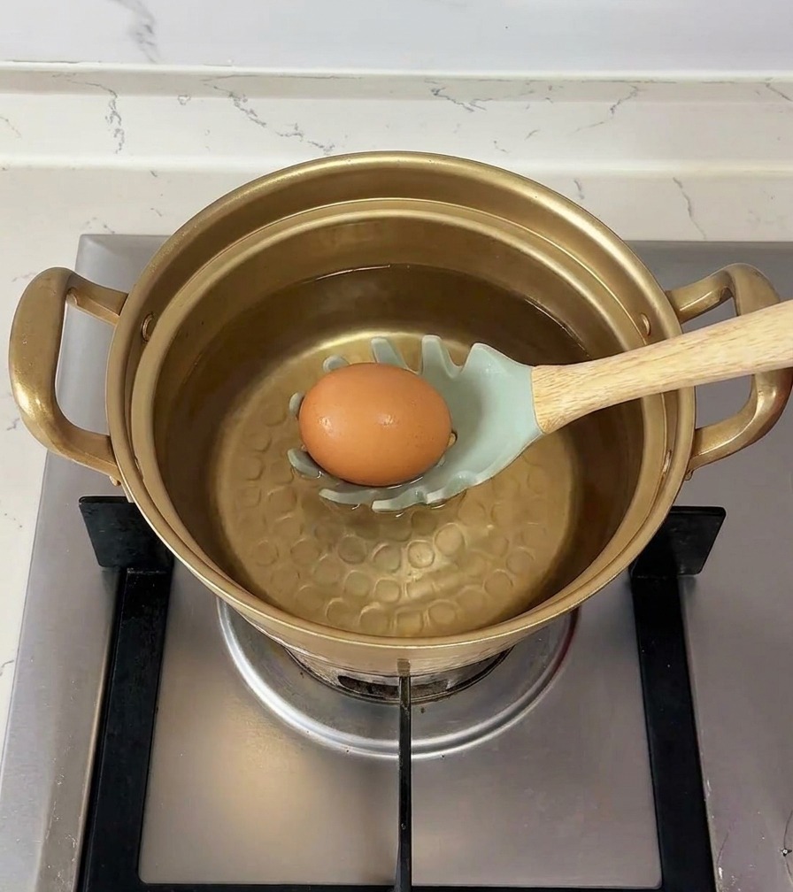 Un œuf brun posé surune écumoire vert clair est délicatement descendu dans une casserole deau.