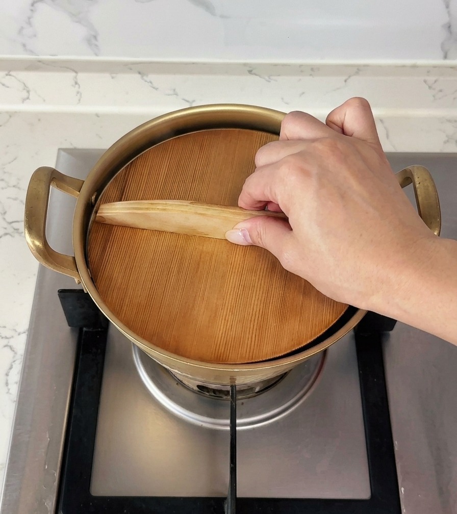 Une main plaçant un couvercle en bois plat sur une petite casserole dorée pour la couvrir.