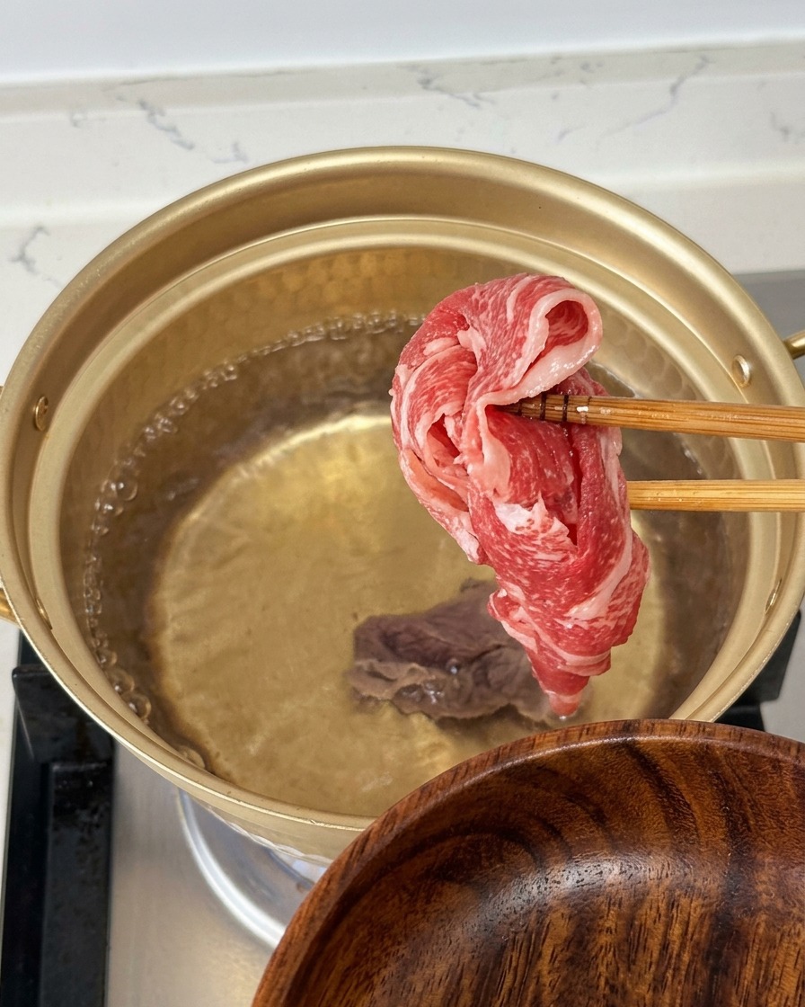 Saisir une fine tranche de bœuf cru et persillé avec des baguettes au-dessus dune casserole dorée deau bouillante.