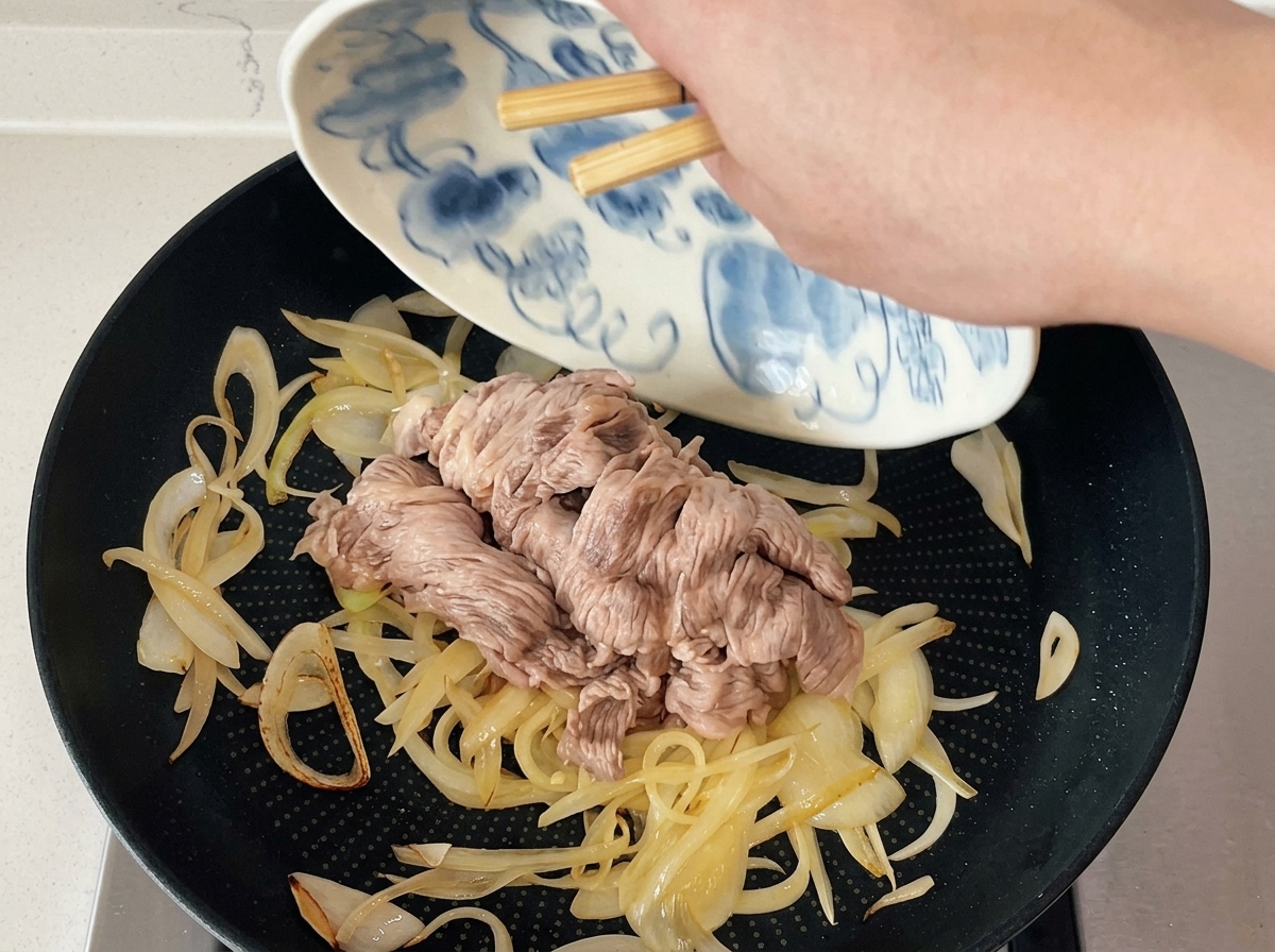 Des baguettes faisant glisser des tranches de bœuf blanchies depuis une assiette à motifs vers une poêle doignons sautés.