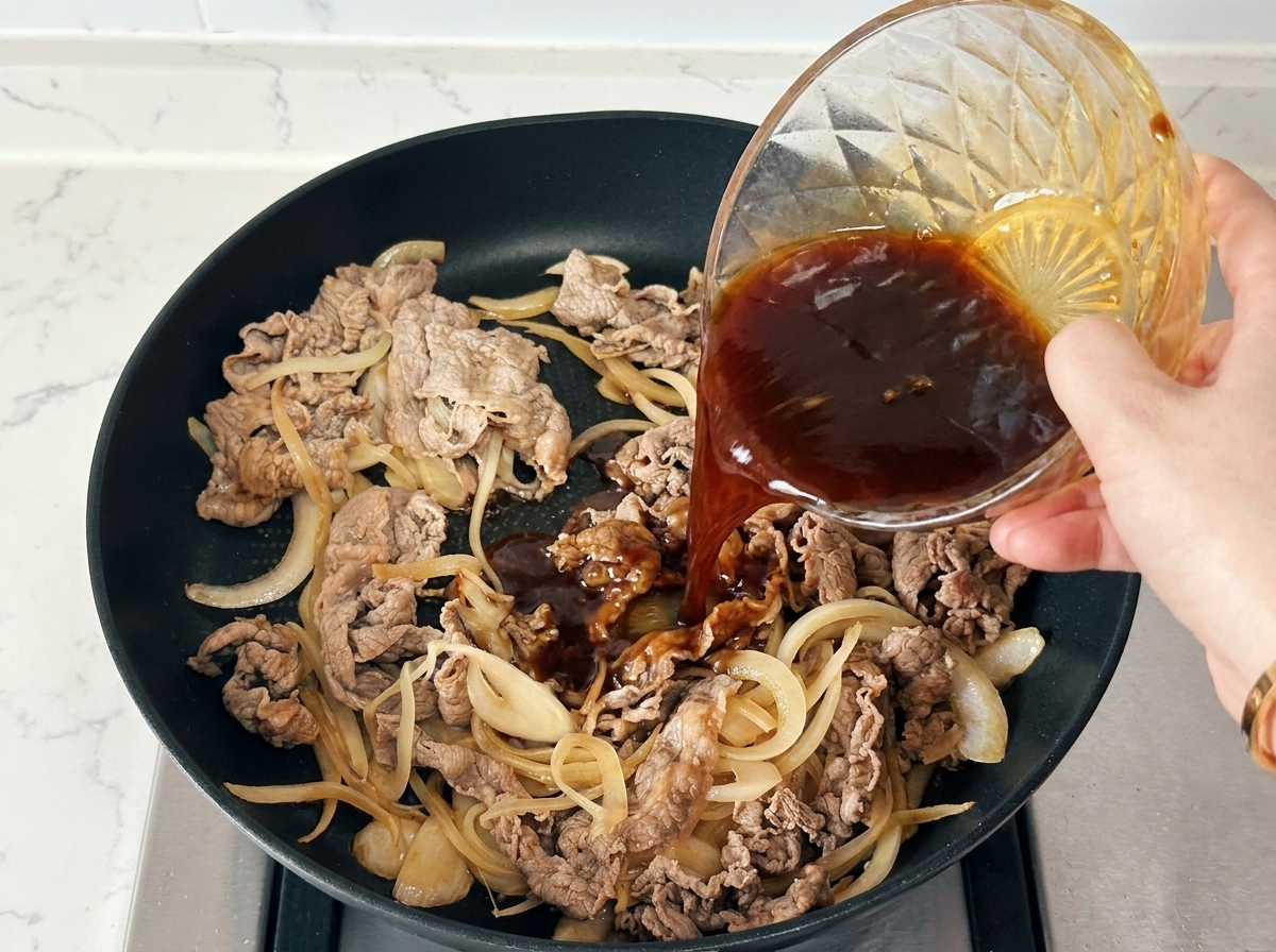 Une main versant une sauce dassaisonnement foncée depuis un bol en verre sur du bœuf et des oignons dans une poêle noire.