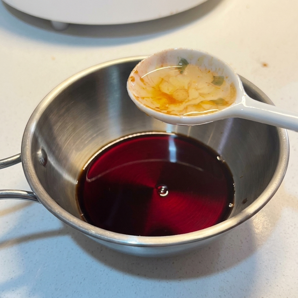 Une cuillère de assaisonnement liquide ajoutée dans un petit bol en métal contenant un mélange de sauce sombre.