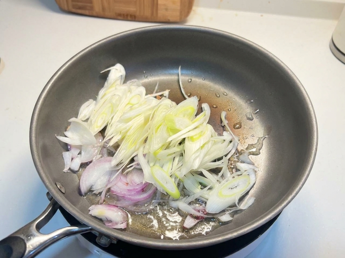 Des oignons blancs émincés et des oignons verts crépitant dans de lhuile chaude au sein dune poêle en métal.