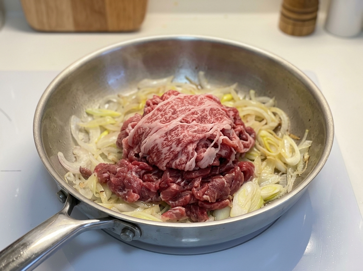 Une portion de bœuf gras cru coupé très finement disposée sur des oignons cuits dans une poêle en métal.