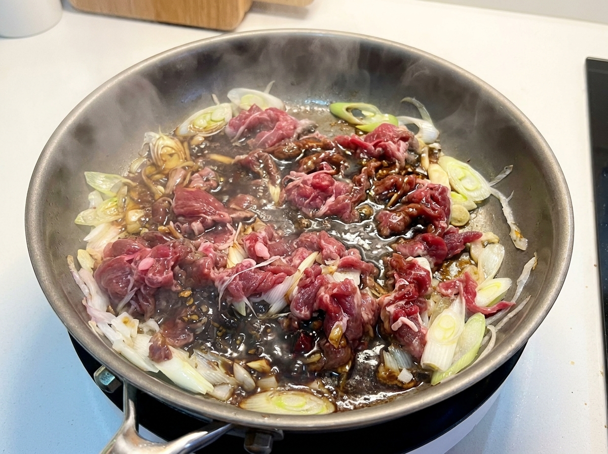 Lamelles de bœuf partiellement cuites et oignons mijotant dans une sauce sombre et riche au sein dune poêle.