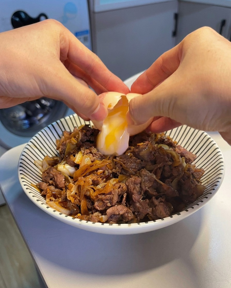 Deux mains écalant un œuf onsen, laissant couler un jaune coulant et un blanc soyeux sur un bol fumant de bœuf et de riz.