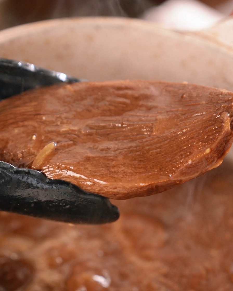 Un morceau épais de langue de bœuf mijotée soulevé dune casserole de sauce sombre avec des pinces de cuisine noires.