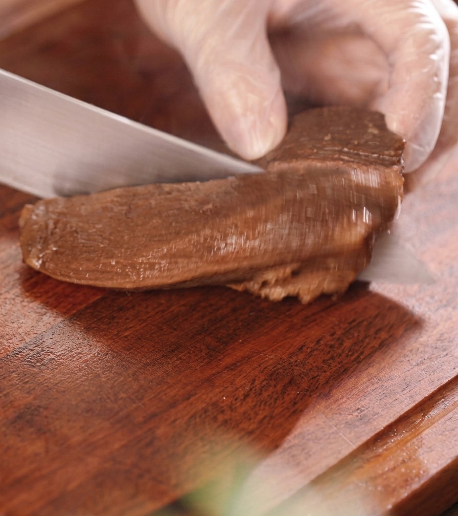 Une main gantée tranchant un morceau de langue de bœuf mijotée en morceaux épais de la taille dune bouchée sur une planche en bois.