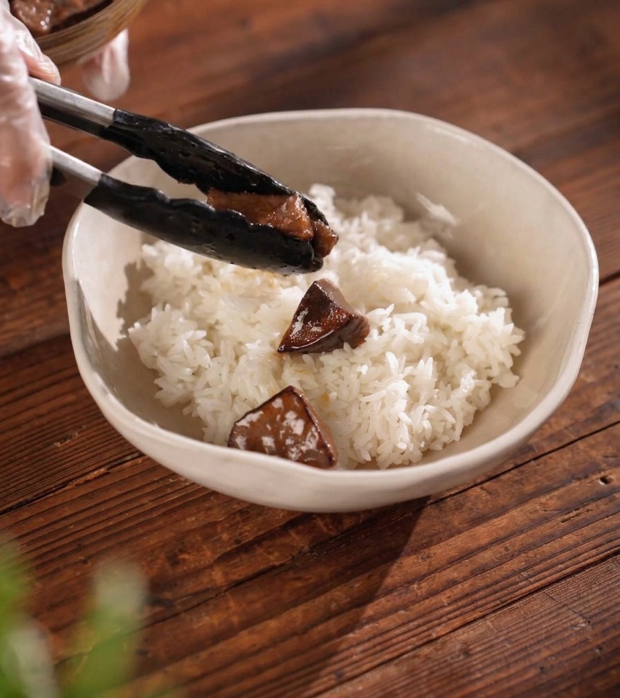 Des pinces déposant un morceau de viande laquée sombre sur un lit de riz blanc bien chaud dans un bol en céramique.