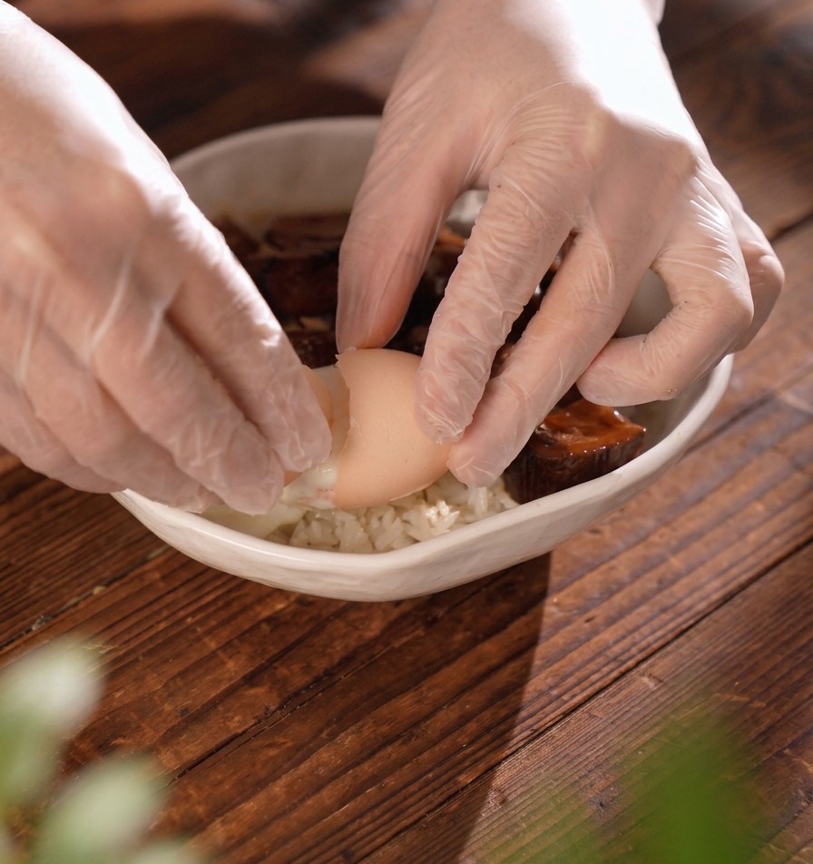 Des mains gantées cassant délicatement une coquille dœuf pour en libérer un œuf poché coulant dans un bol de riz à la viande.
